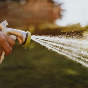 A close-up photograph of hands holding a garden hose nozzle spraying a fine mist of water, highlighting the importance of fall winterization to prevent frozen outdoor pipes.